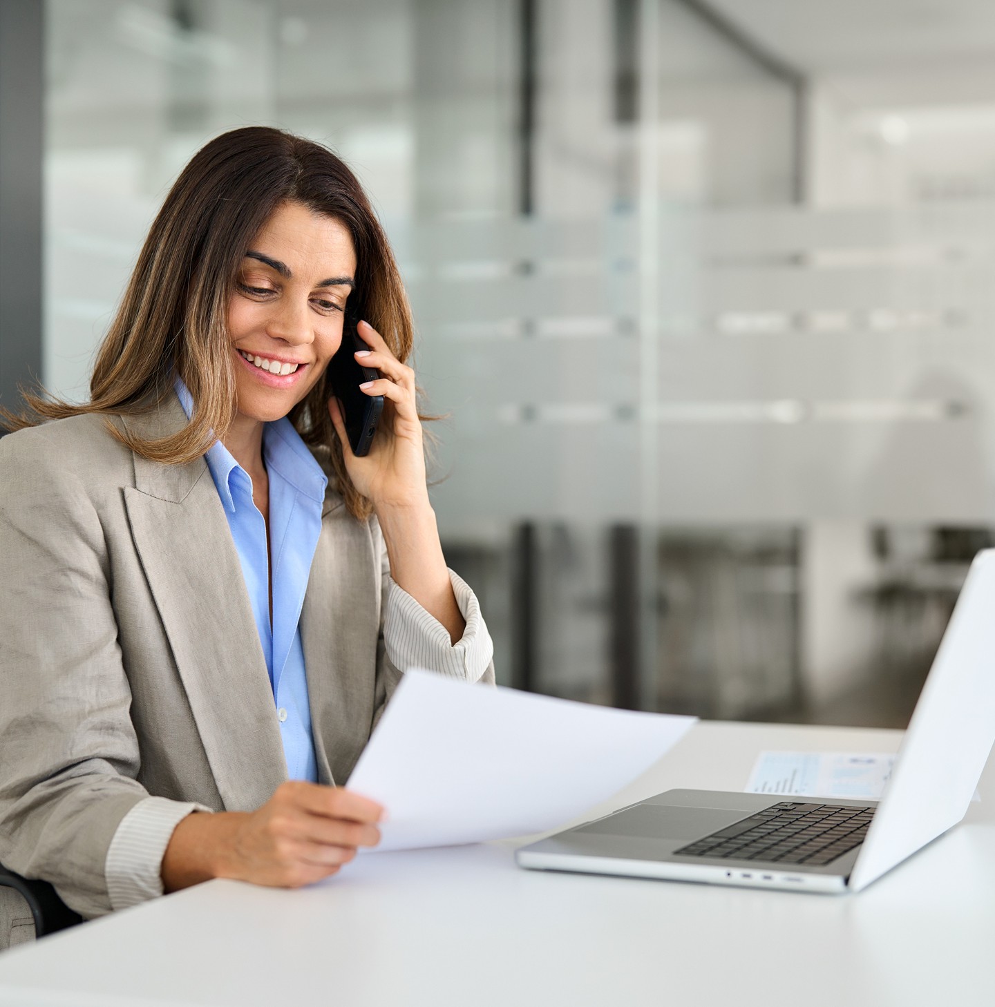 A woman in a beige suit holds papers while talking on the phone, sitting at a desk with a laptop in a modern office setting.