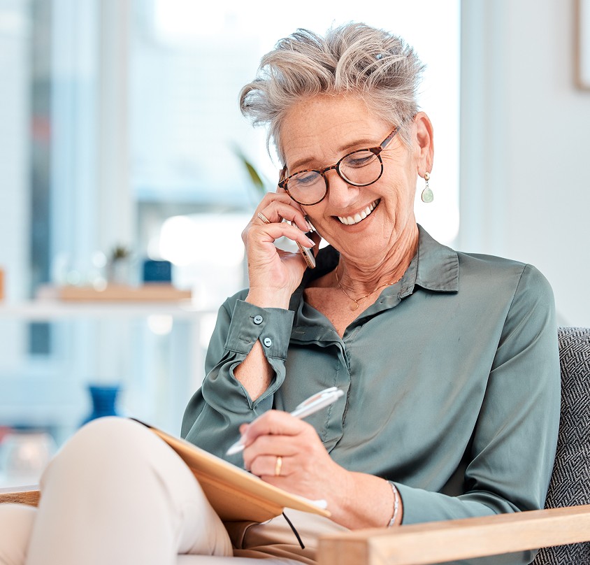 An elderly woman sits thoughtfully with a notebook and pen, wearing a light green shirt and holding a phone in a bright room.